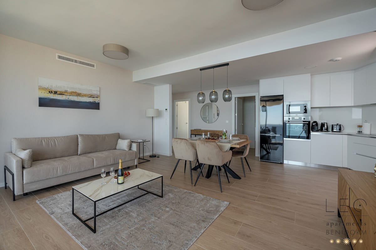 Uma sala de estar moderna com sofá, mesa de centro e área de jantar. Há uma cozinha com armários e eletrodomésticos brancos.