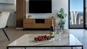 A modern living room with a marble coffee table, a bottle of champagne, and a glass. A large flat-screen TV is mounted on the wall, and there is a potted plant to the right. - Torre Sakura (Benidorm)