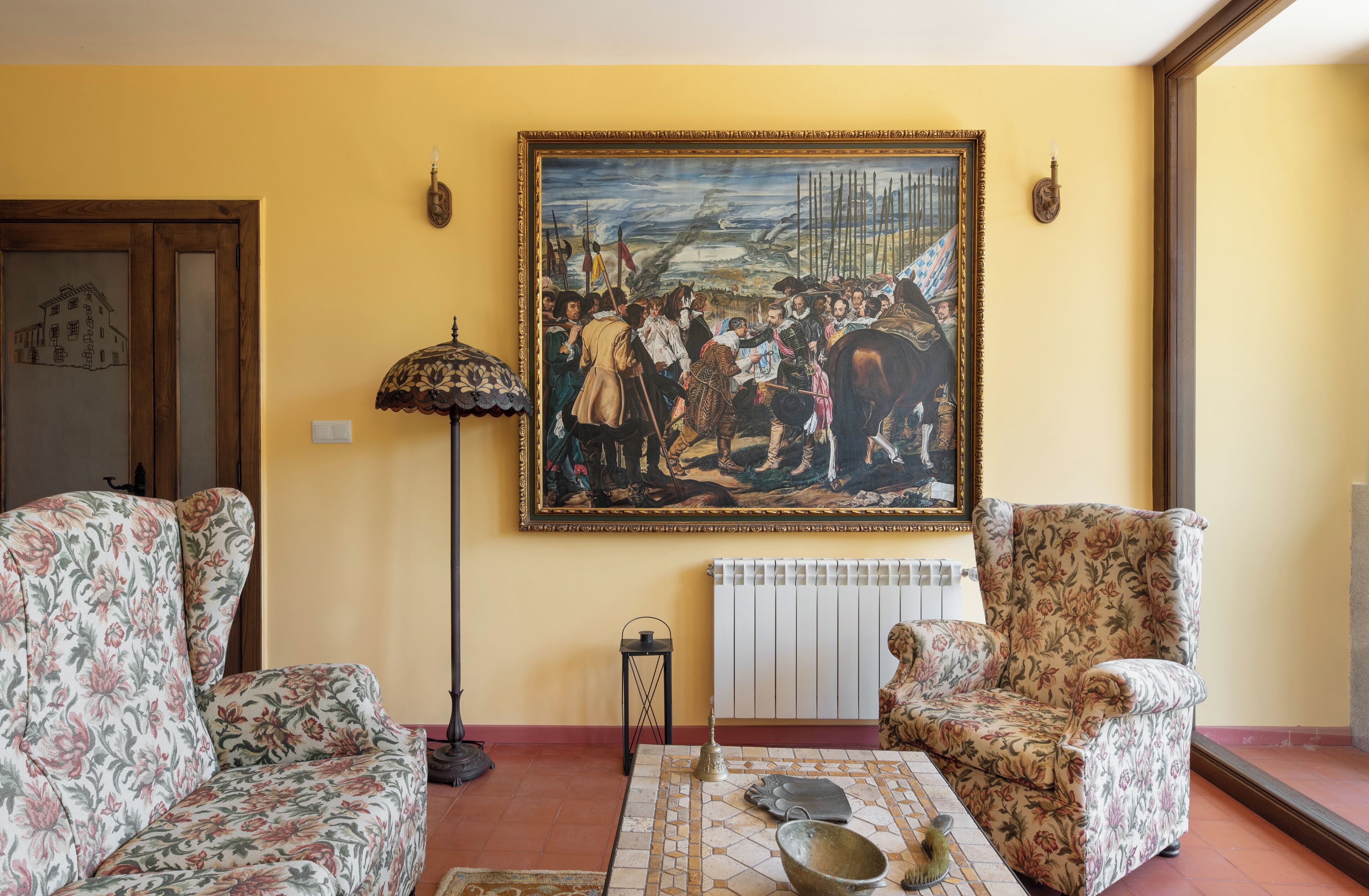 A living room with floral-patterned armchairs, a table with a bowl and gloves, a painting on the wall, and a radiator.