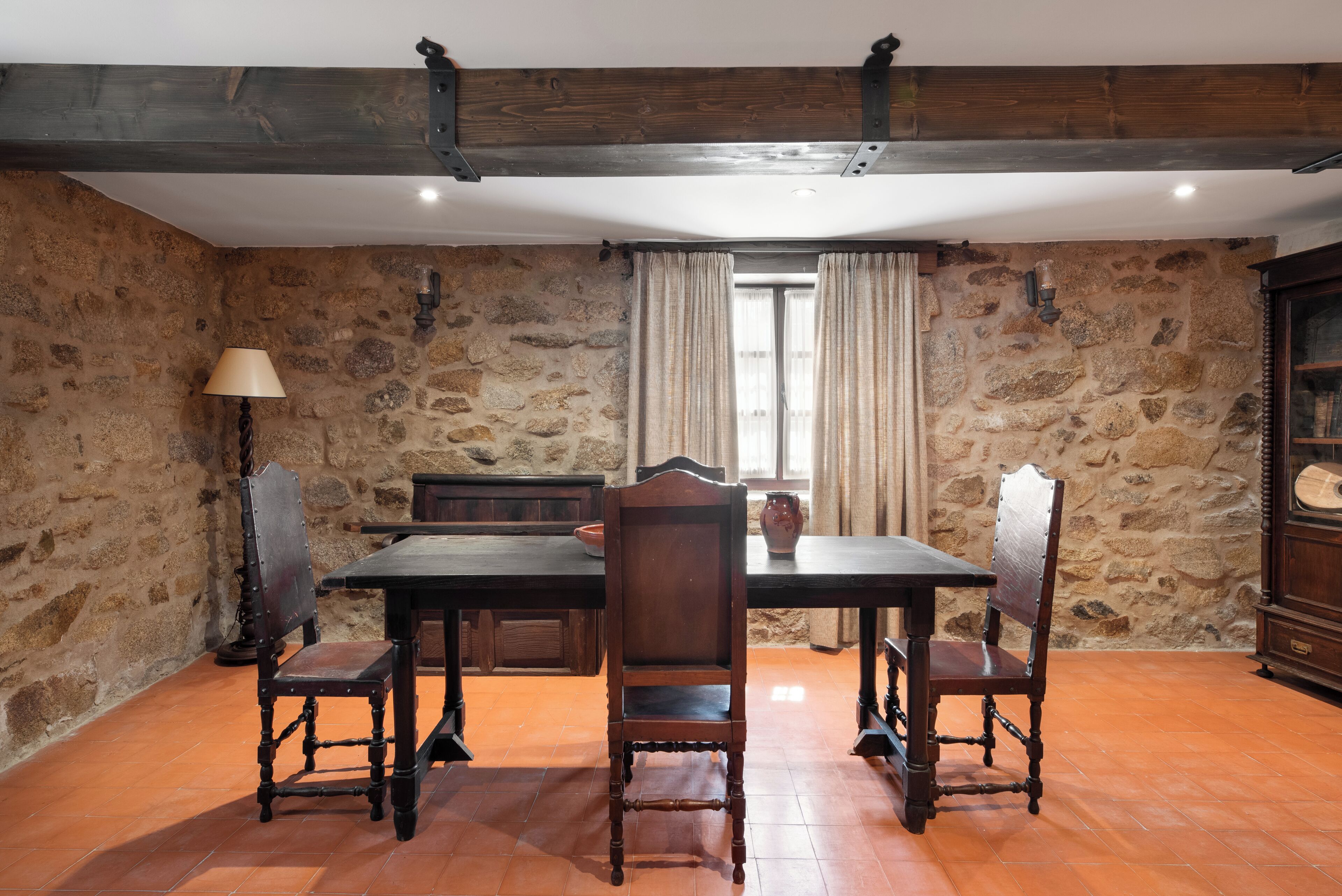 A rustic dining area with a stone wall, wooden table, and antique chairs.