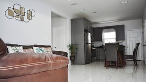 A modern living room with a brown leather sofa, a dining area with a table and chairs, a kitchen with a refrigerator, and a potted plant.