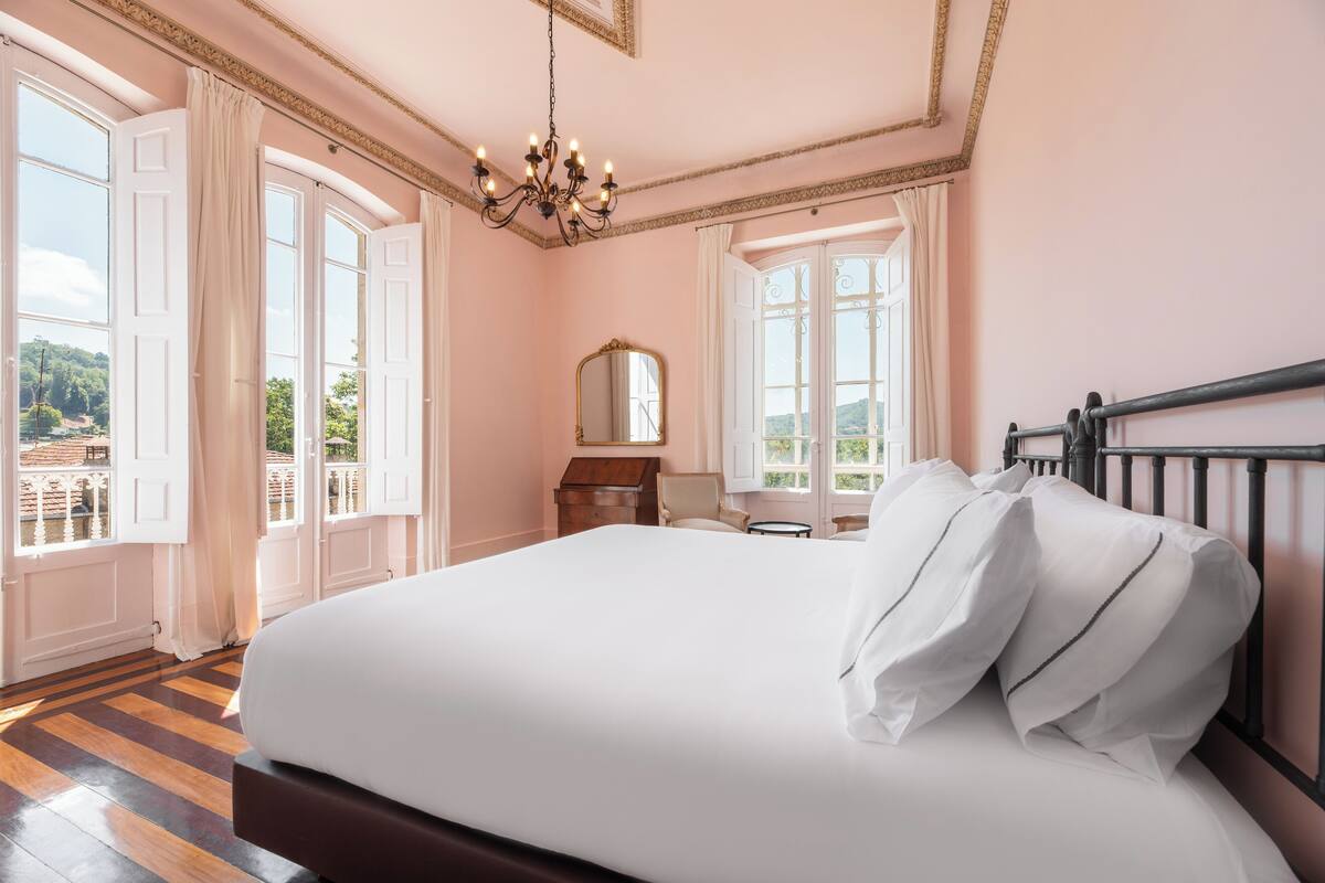 Ein Schlafzimmer mit einem großen Bett, einem Kronleuchter, einem hölzernen Schminktisch und Blick ins Freie durch große Fenster.