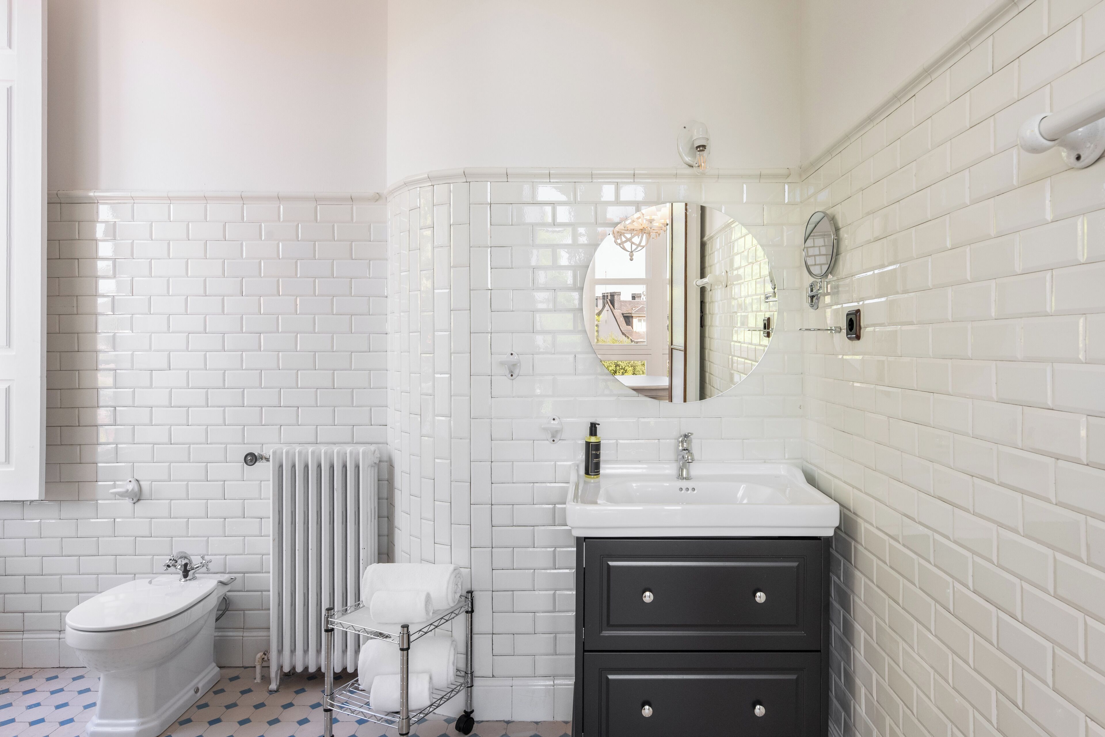 Une salle de bain avec des carreaux de céramique blancs rectangulaires, des toilettes blanches, un lavabo blanc avec un miroir et un meuble-lavabo noir.