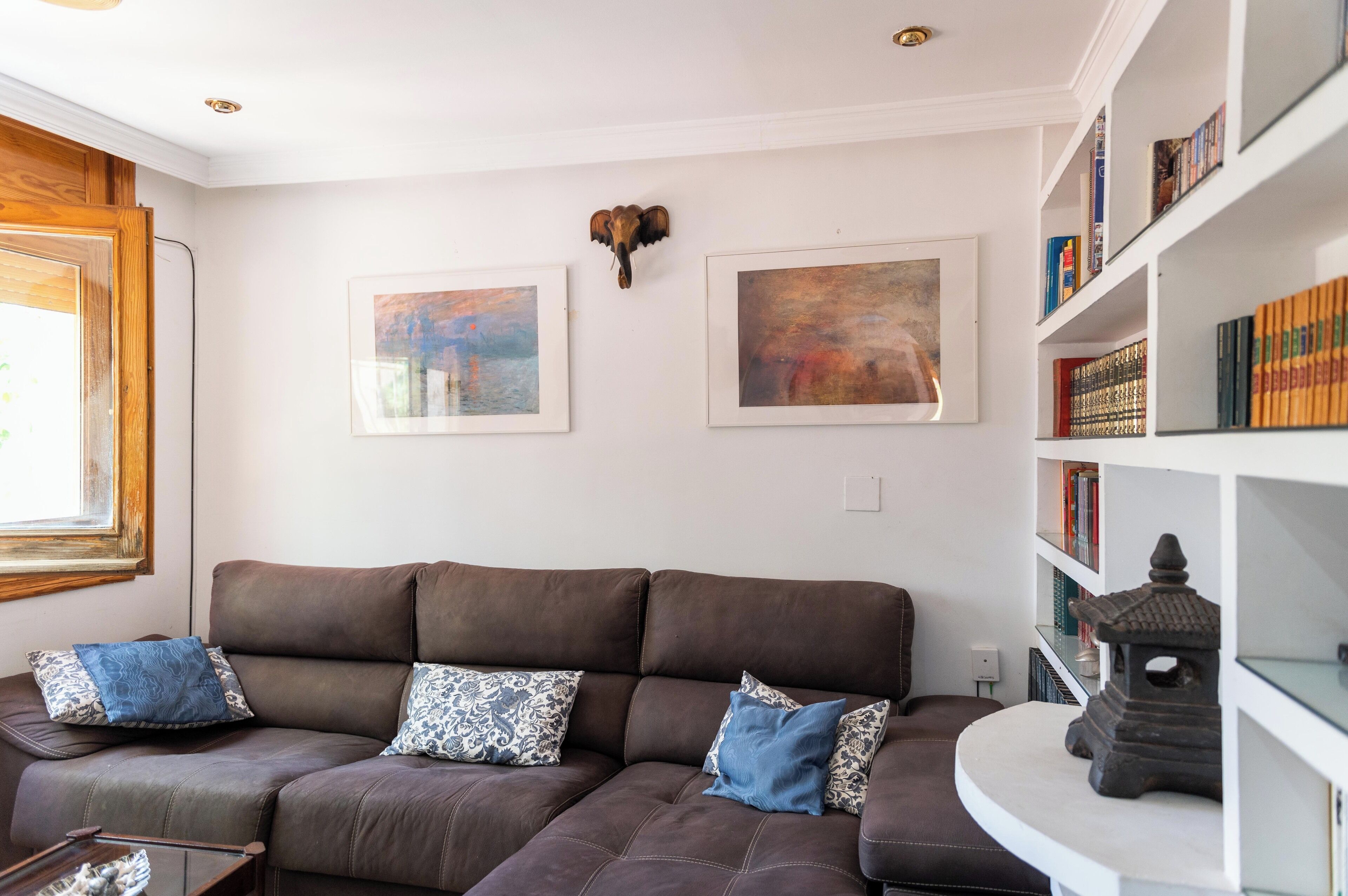 A living room with a brown sofa, two framed paintings, a bookshelf, and a decorative wall piece.