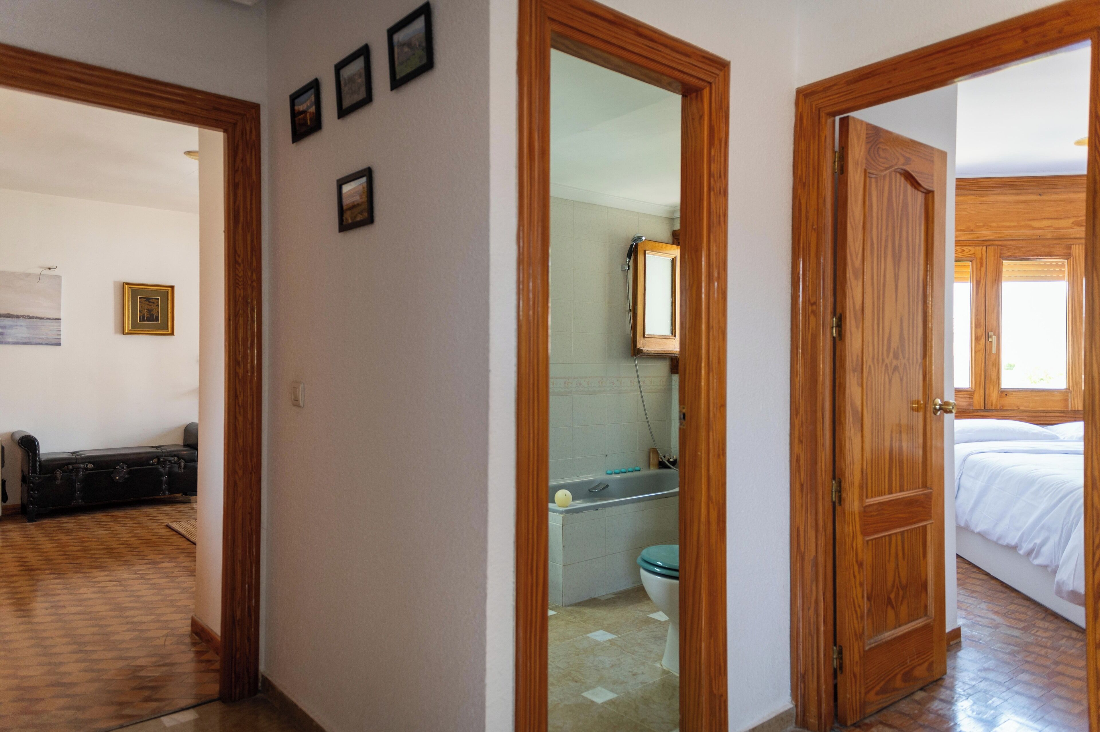A hallway leading to a bathroom with a toilet, a mirror, and a shower. There is a bed visible in the adjacent room.