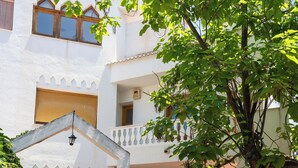 A white building with multiple windows and balconies, a wooden bench, and a tree in the foreground. - Piscina, gym,2 barbacoas, vistas a Sierra Nevada desde terraza en ático de 60m2  (Peligros)