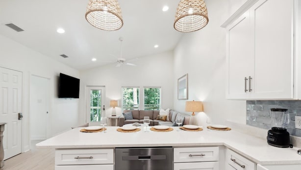 Une cuisine moderne avec des meubles blancs, un grand îlot central doté d’un lave-vaisselle, d’un four encastré et d’un mixeur noir. On y trouve deux lustres suspendus en osier, un ventilateur de plafond et un téléviseur à écran plat fixé au mur.