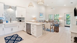 Eine moderne Küche mit weißen Schränken, einem Geschirrspüler und einer zentralen Kochinsel. Ein Essbereich mit Tisch und Stühlen, einer Couch und einem Deckenventilator.