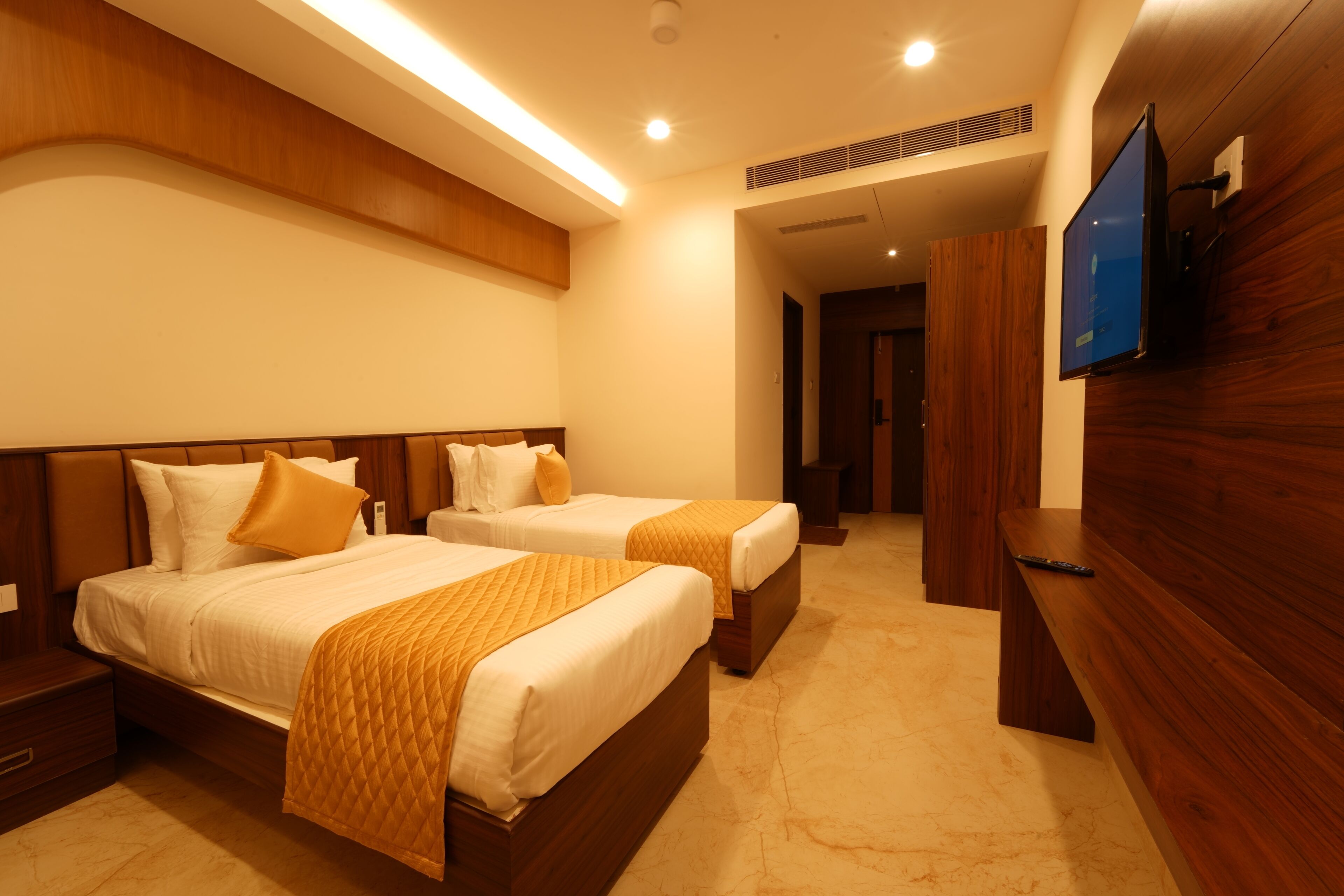 Habitación de hotel con dos camas, cabeceros de madera, un televisor de pantalla plana montado en la pared y un banco junto a la ventana.