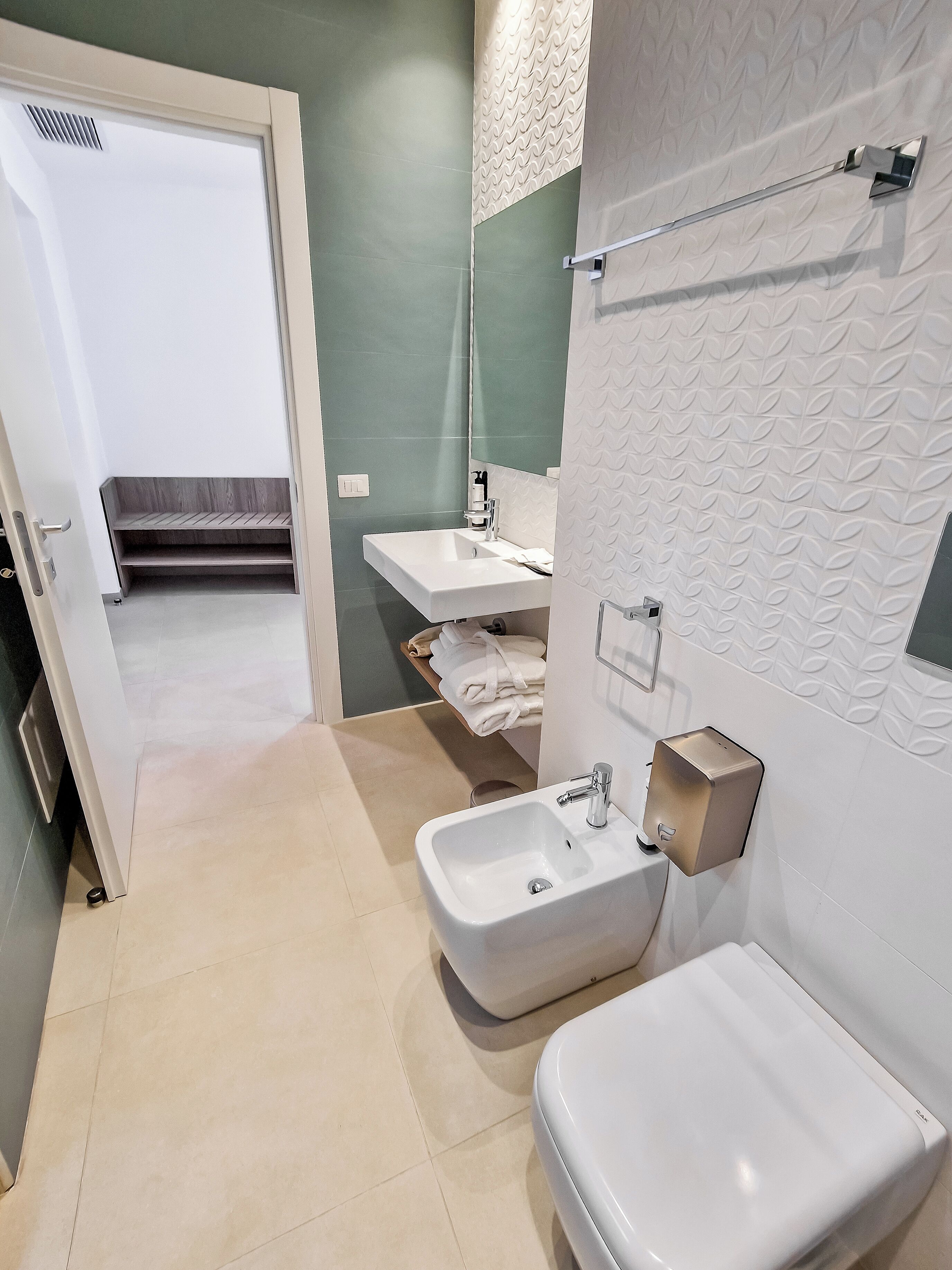 A modern bathroom with a white sink, a toilet, and a mirror. There is a towel rack on the wall and a shelf with towels.