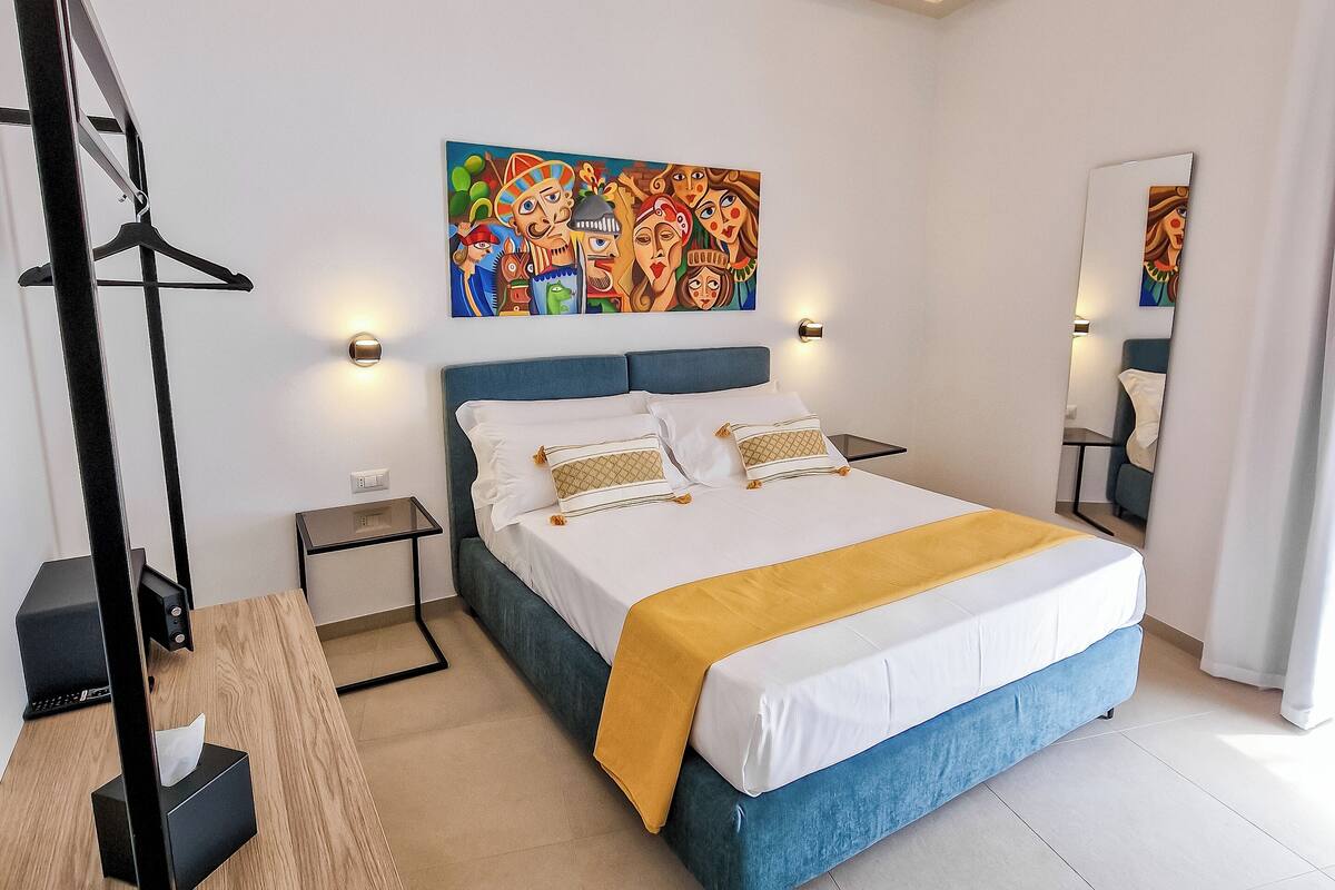 Une chambre à coucher moderne avec un grand lit, des tables de chevet, un porte-manteau mural et un tableau abstrait aux couleurs vives accroché au mur.