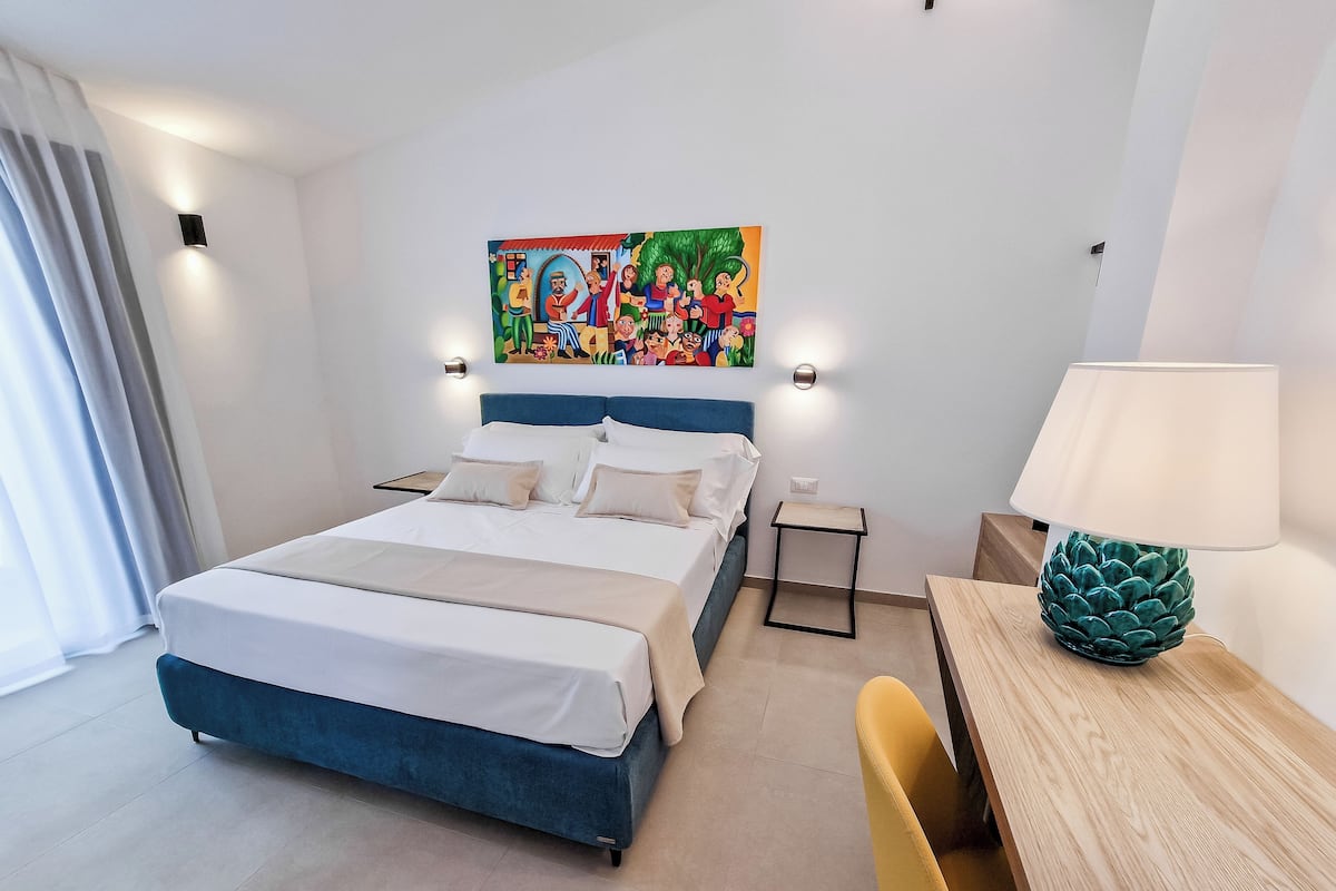 Une chambre d’hôtel avec un lit, des tables de chevet, un bureau, une chaise, une lampe et un tableau coloré au mur.