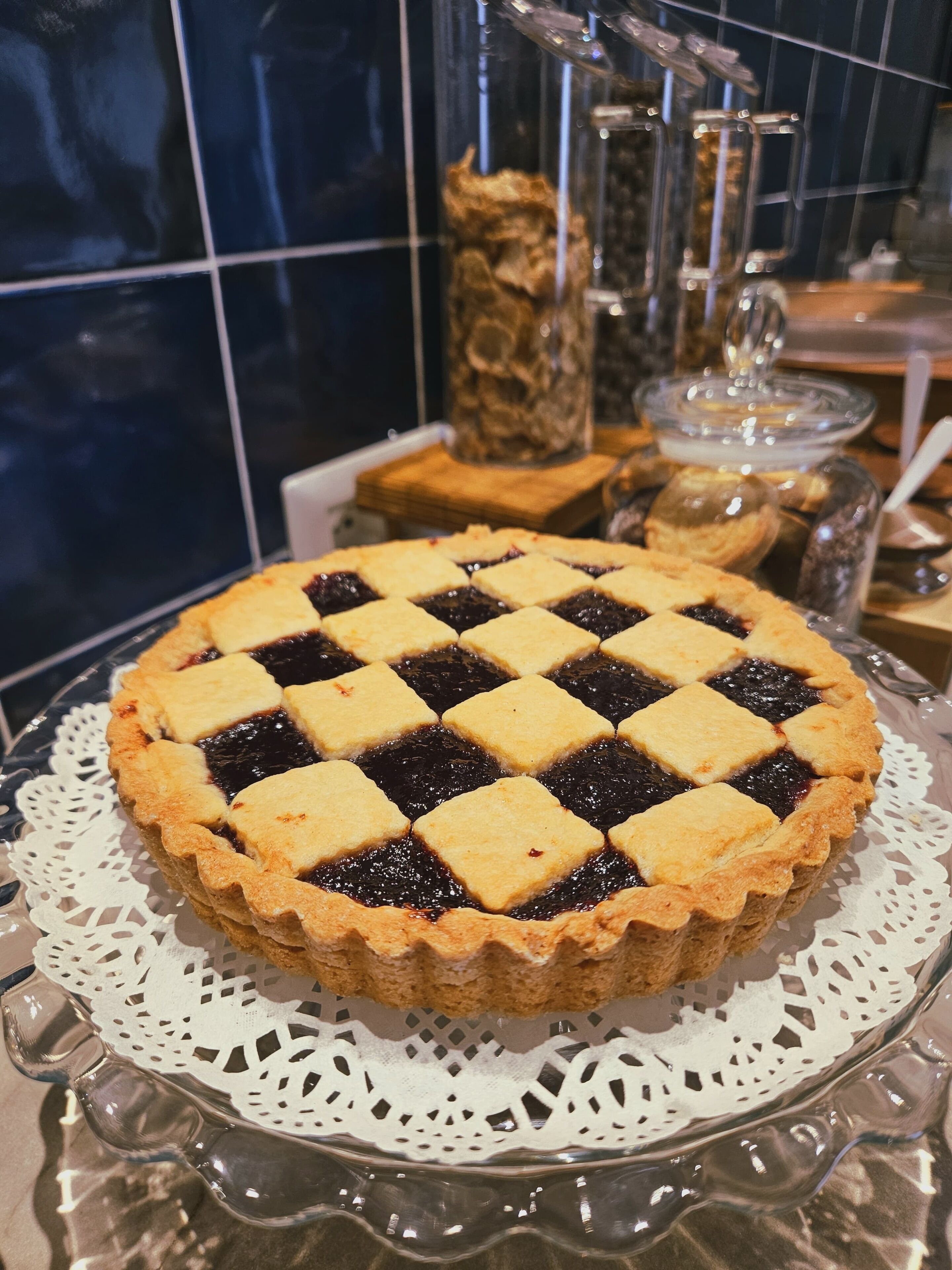 Une tarte recouverte d’une pâte en croisillons, présentée sur une assiette en verre ornée d’un chemin de table, le tout posé sur une table dont le mur est recouvert de carreaux.