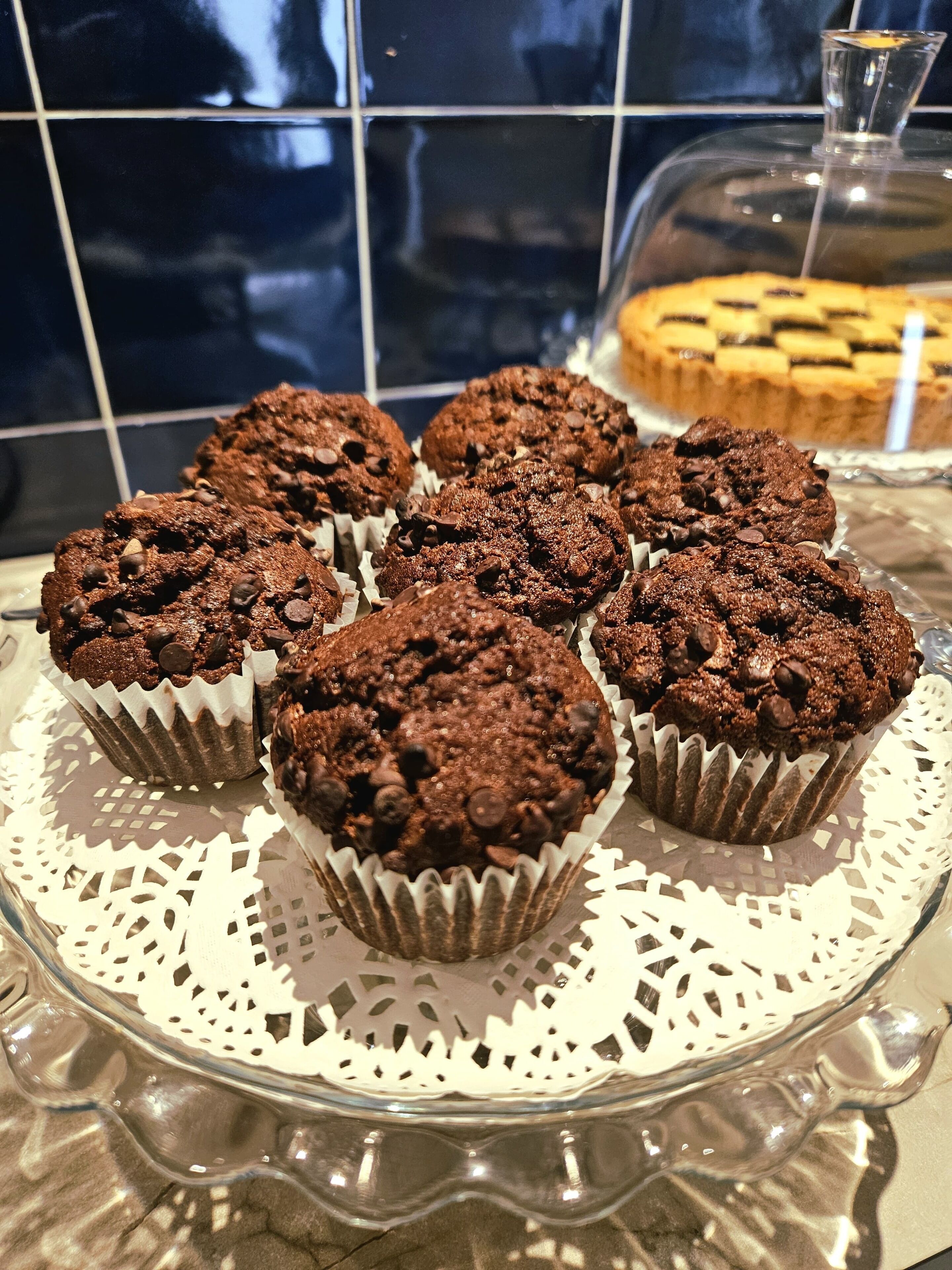 Bandeja de muffins de chocolate sobre un plato de vidrio con un mantel de encaje, en una cocina con salpicadero de azulejos.