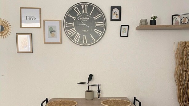 Un coin repas avec une table en bois et deux chaises, une grande horloge murale et plusieurs tableaux encadrés.