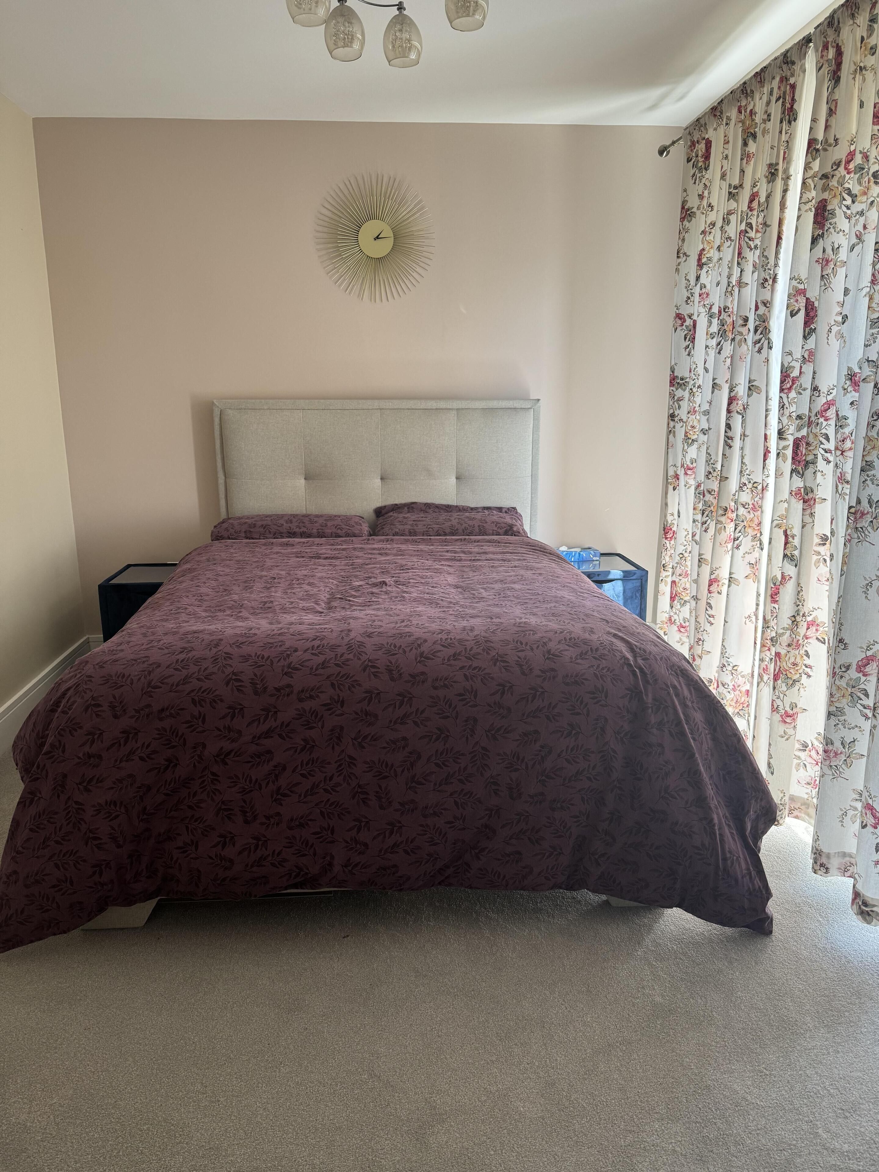 A bedroom with a large bed, a nightstand, a wall clock, and a window with floral curtains.