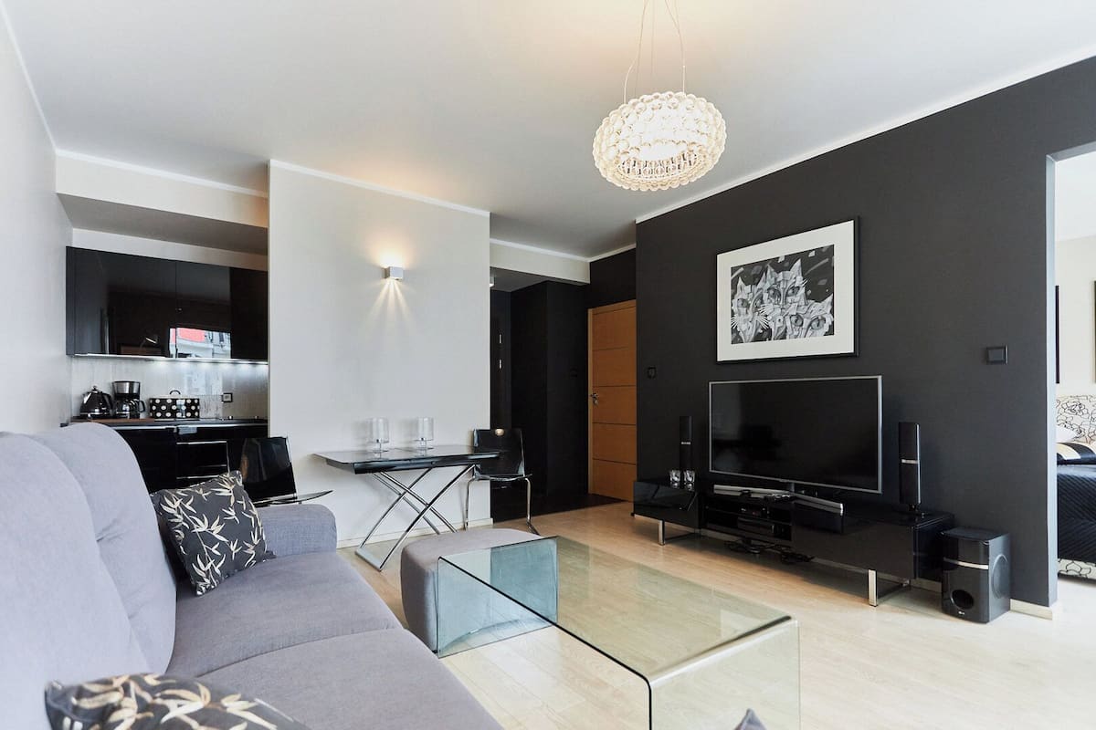 A modern living room with a grey sofa, a glass coffee table, a flat-screen TV, and a dining area with a small table and chairs.