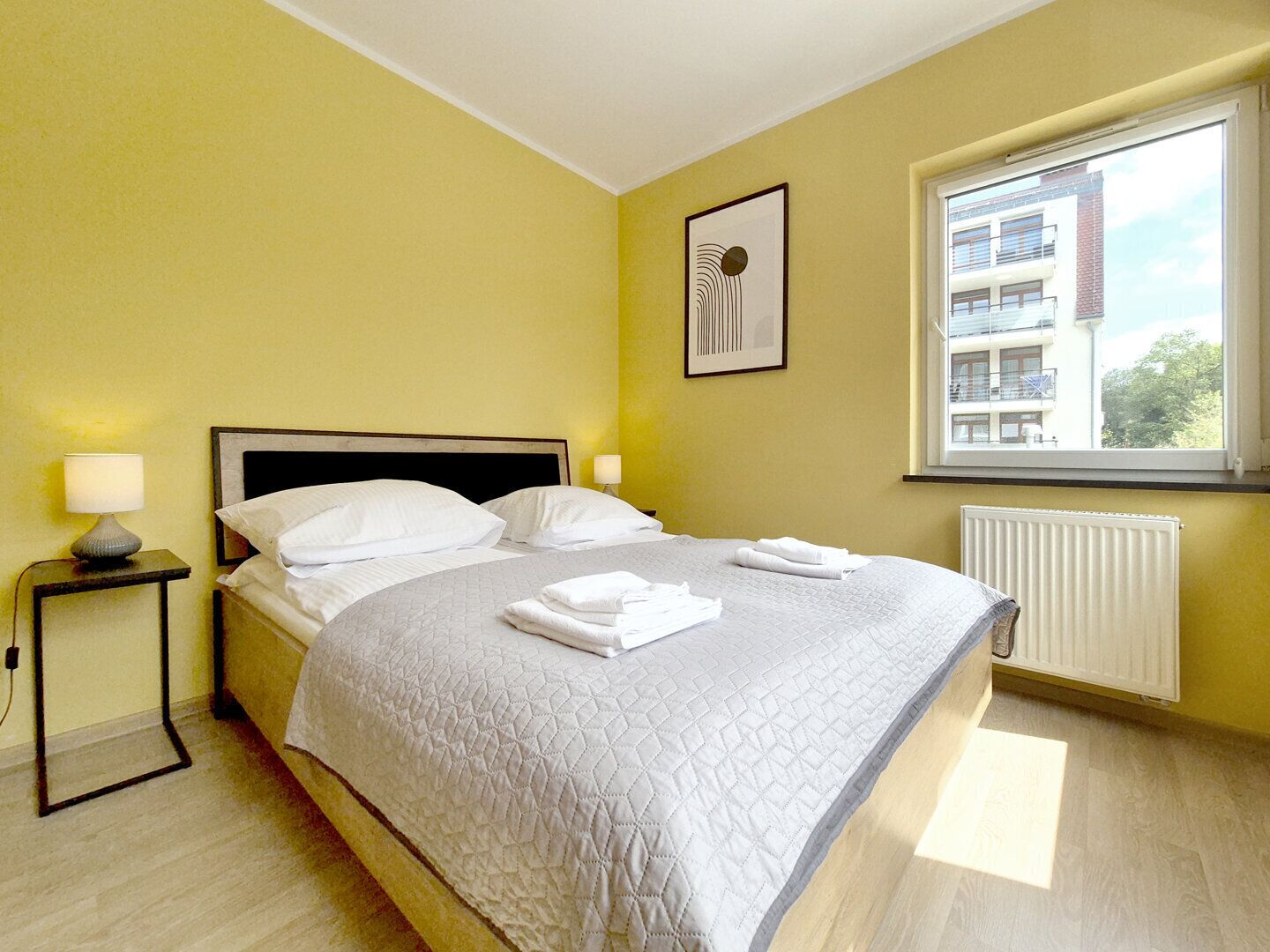 Habitación de hotel con cama, dos lámparas de noche, un cuadro en la pared, ventana con vistas a edificios y un radiador.