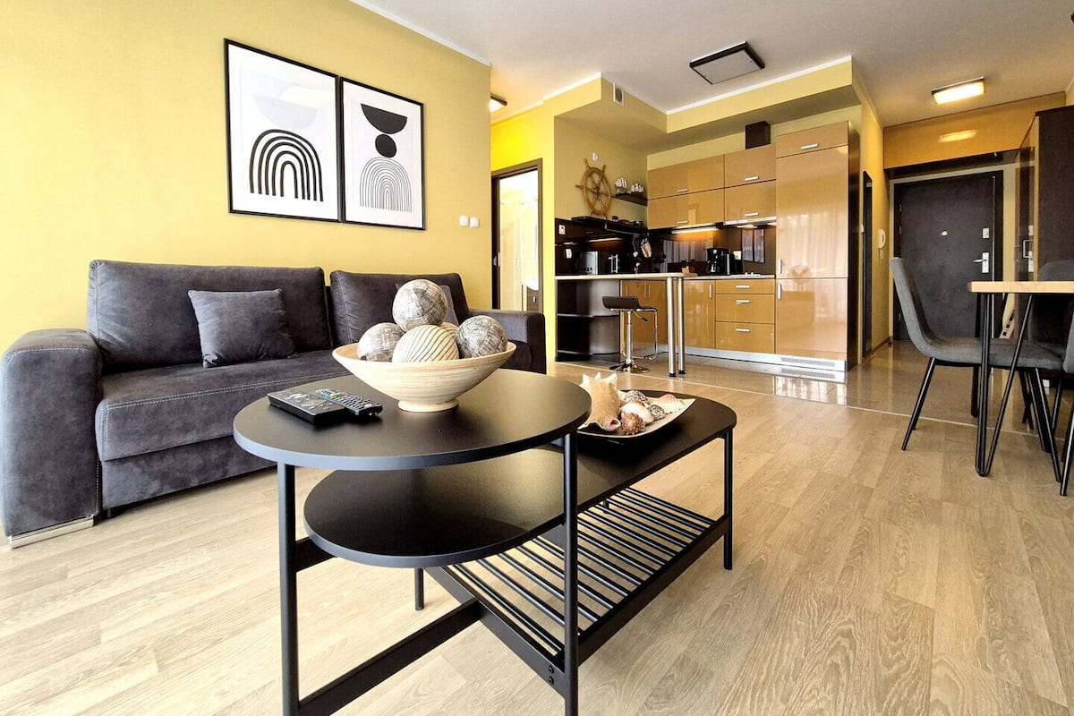 A modern living room with a sofa, a coffee table with a bowl of decorative balls, a dining area with a table and chairs, and a kitchen in the background.
