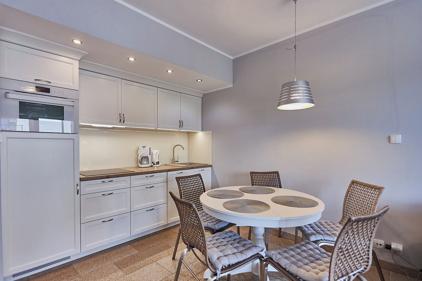 A compact kitchen with white cabinets, a built-in oven, and a coffee maker. There is a round dining table with four chairs, and a hanging pendant light above.