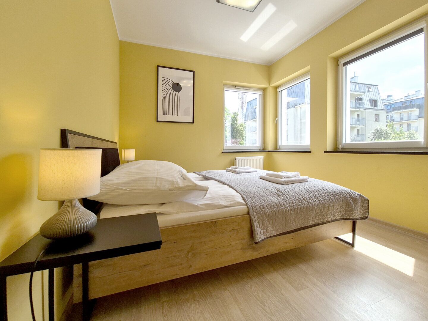 A modern bedroom with a wooden bed, a bedside table with a lamp, and two windows providing views of buildings.