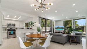 Eine moderne Küche mit weißen Schränken, ein Essbereich mit einem runden Tisch und weißen Stühlen, ein graues Sofa und ein großes Fenster mit Blick auf einen Pool.