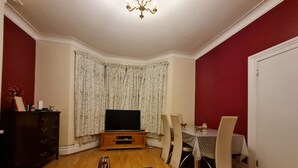 A living room with a TV on a wooden stand, a dining table with chairs, a glass coffee table, and a red wall. - House-sleeps4-parking-garden (London)