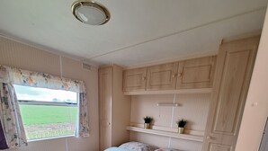 A bedroom with a bed, a window with curtains, a ceiling light, and wooden cabinets. - Gezellige Ruime Stacaravan om Lekker tot Rust te Komen bij ons op de Camping (Burum)