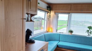 A compact RV interior with a kitchenette, a seating area with a cushioned bench, and large windows offering a view of the outdoors. - Gezellige Ruime Stacaravan om Lekker tot Rust te Komen bij ons op de Camping (Burum)