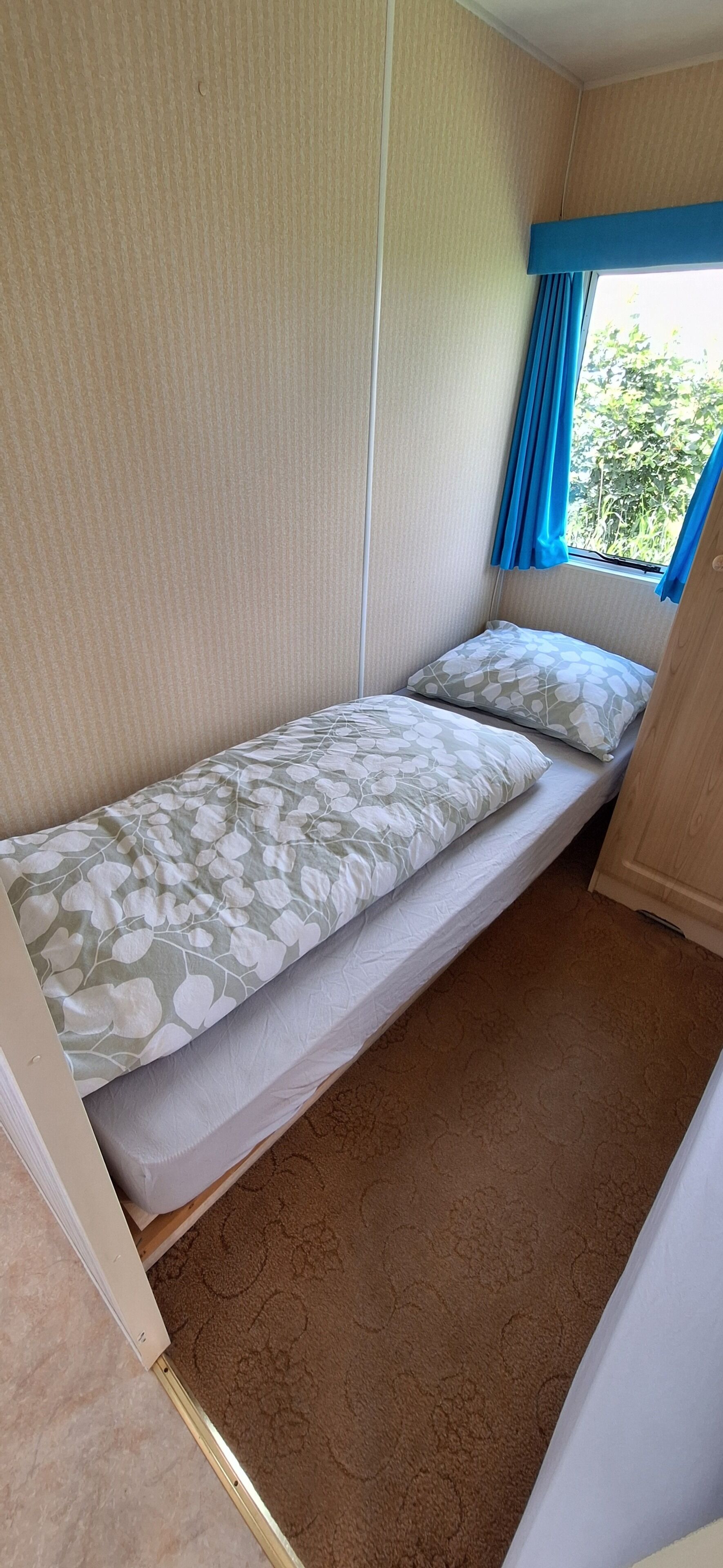 A compact room with a single bed, a window with blue curtains, and a beige wall.