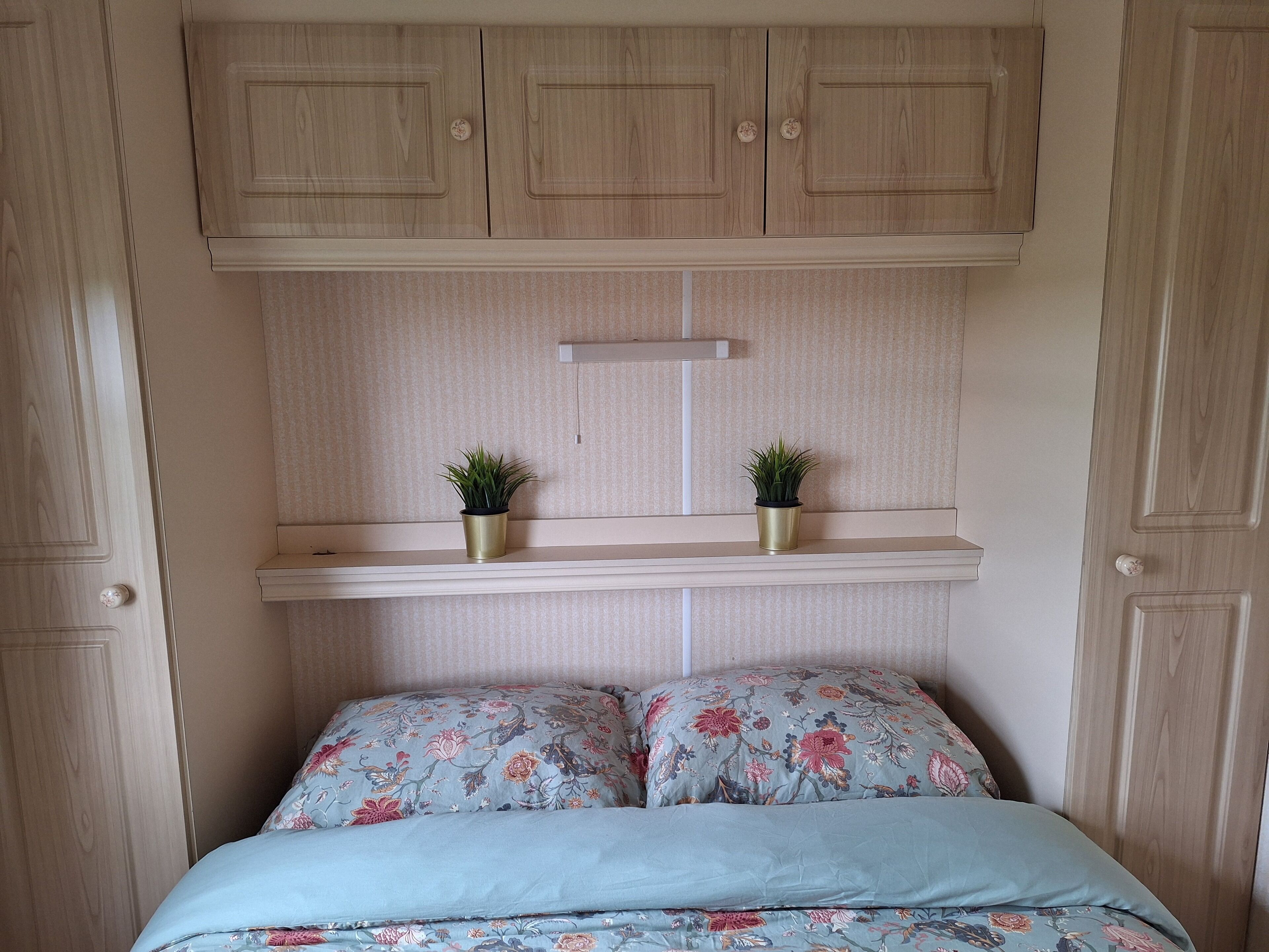 Ein Schlafzimmer mit einem Bett, floralem Bettzeug, zwei Blumentöpfen auf Regalen und Holzschränken darüber.