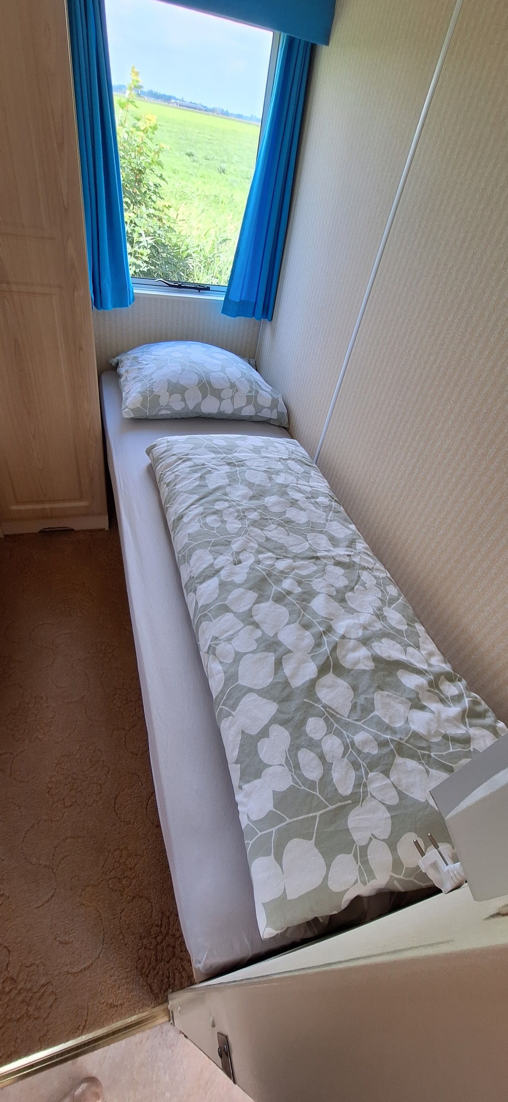 Habitación compacta con una cama individual, ventana con cortinas y vista a un campo verde.