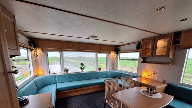 A cozy RV interior with a blue cushioned bench, a dining table, and a kitchen area with wooden cabinets. - Gezellige Ruime Stacaravan om Lekker tot Rust te Komen bij ons op de Camping (Burum)