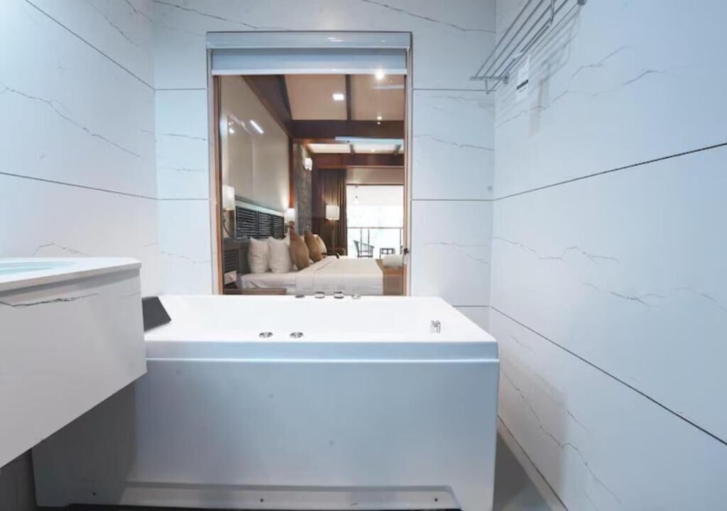 A modern bathroom with white marble walls, a large bathtub, and a visible bedroom area with a bed and curtains.