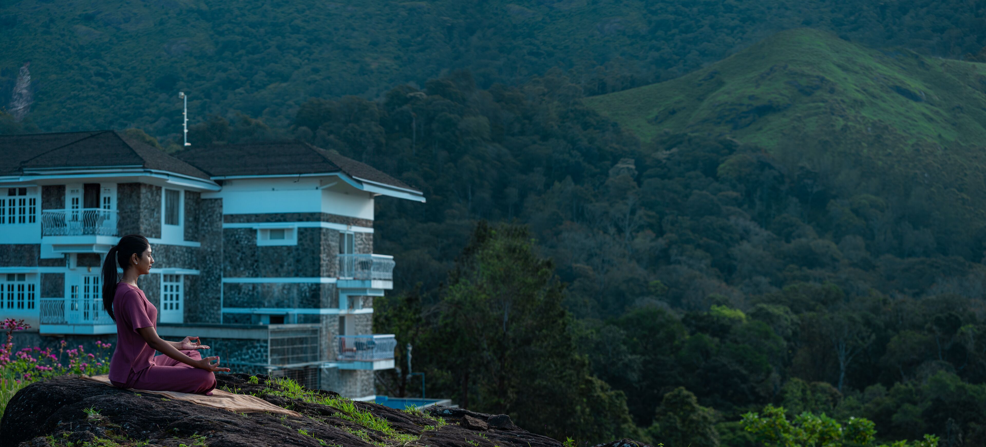 A person meditating on a rock with a large house in the background.