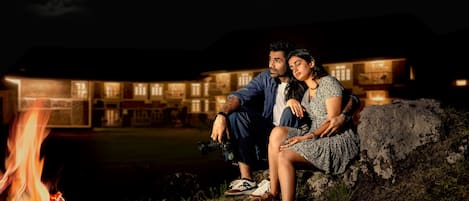 A couple sitting by a campfire at night with a lit building in the background.
