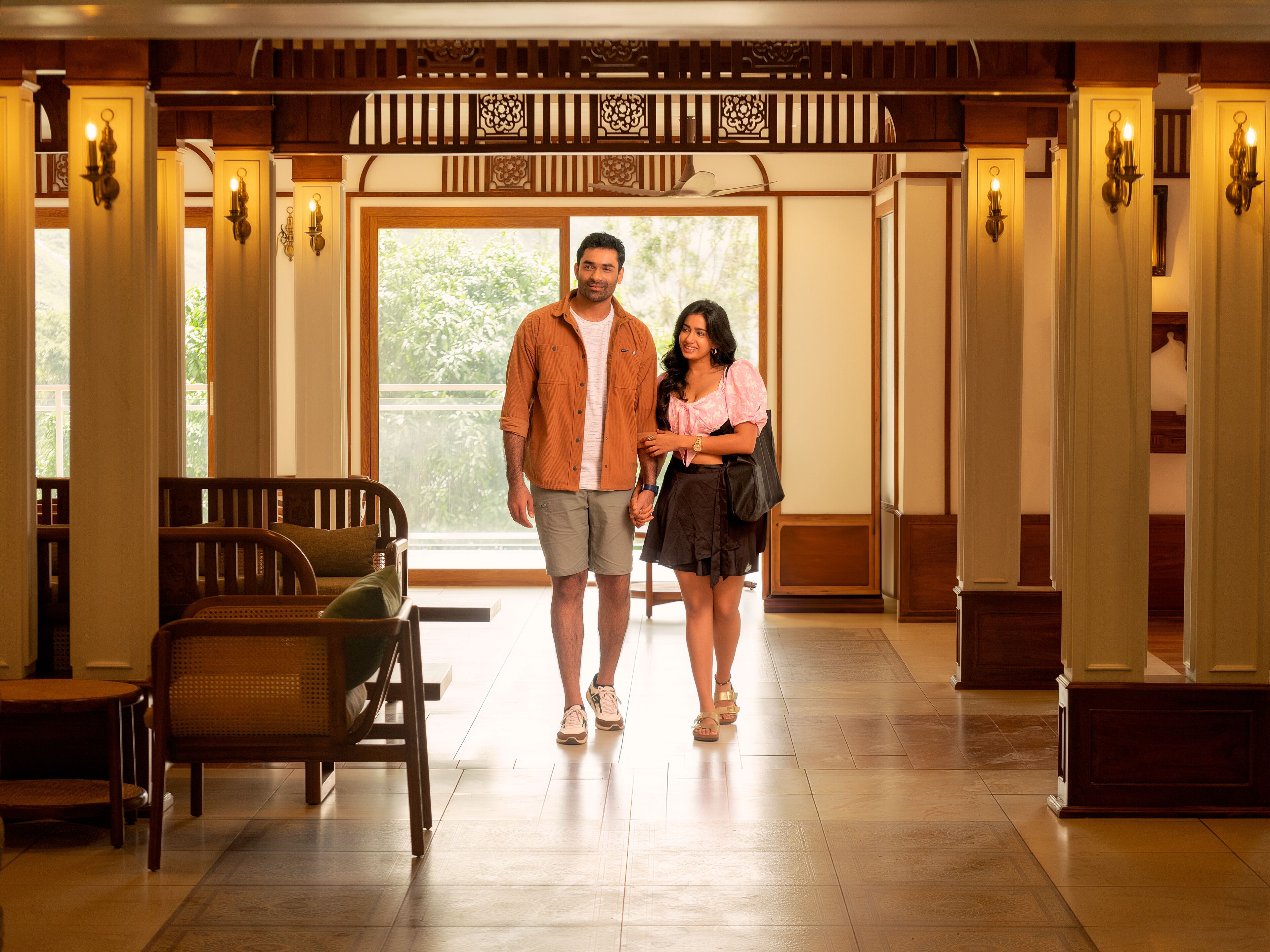 A man and woman walking through a lobby with wooden paneling, a bench, and a large window with a view of greenery.