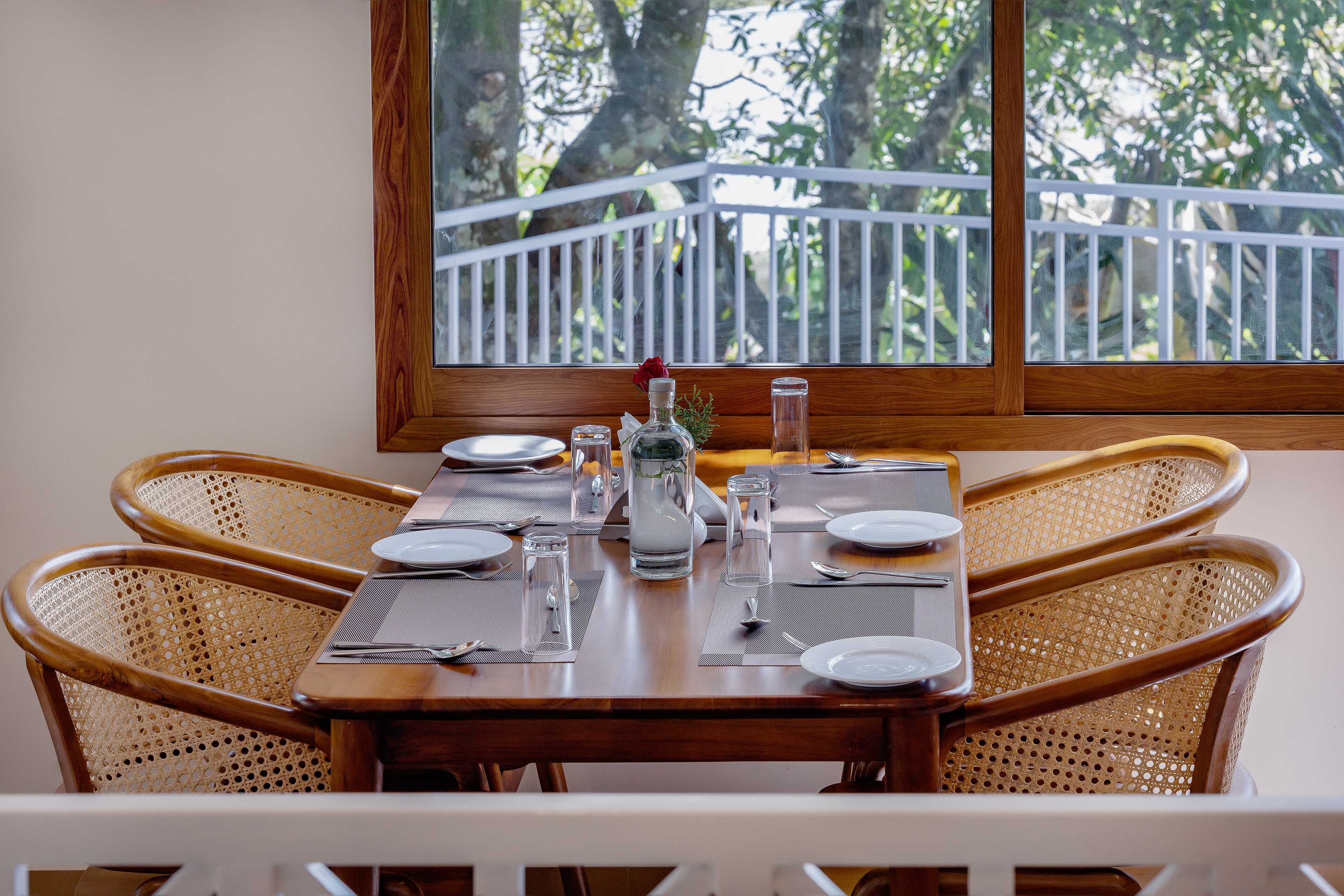 A dining table set with plates, glasses, and cutlery, surrounded by wicker chairs, with a view of trees through a balcony railing.