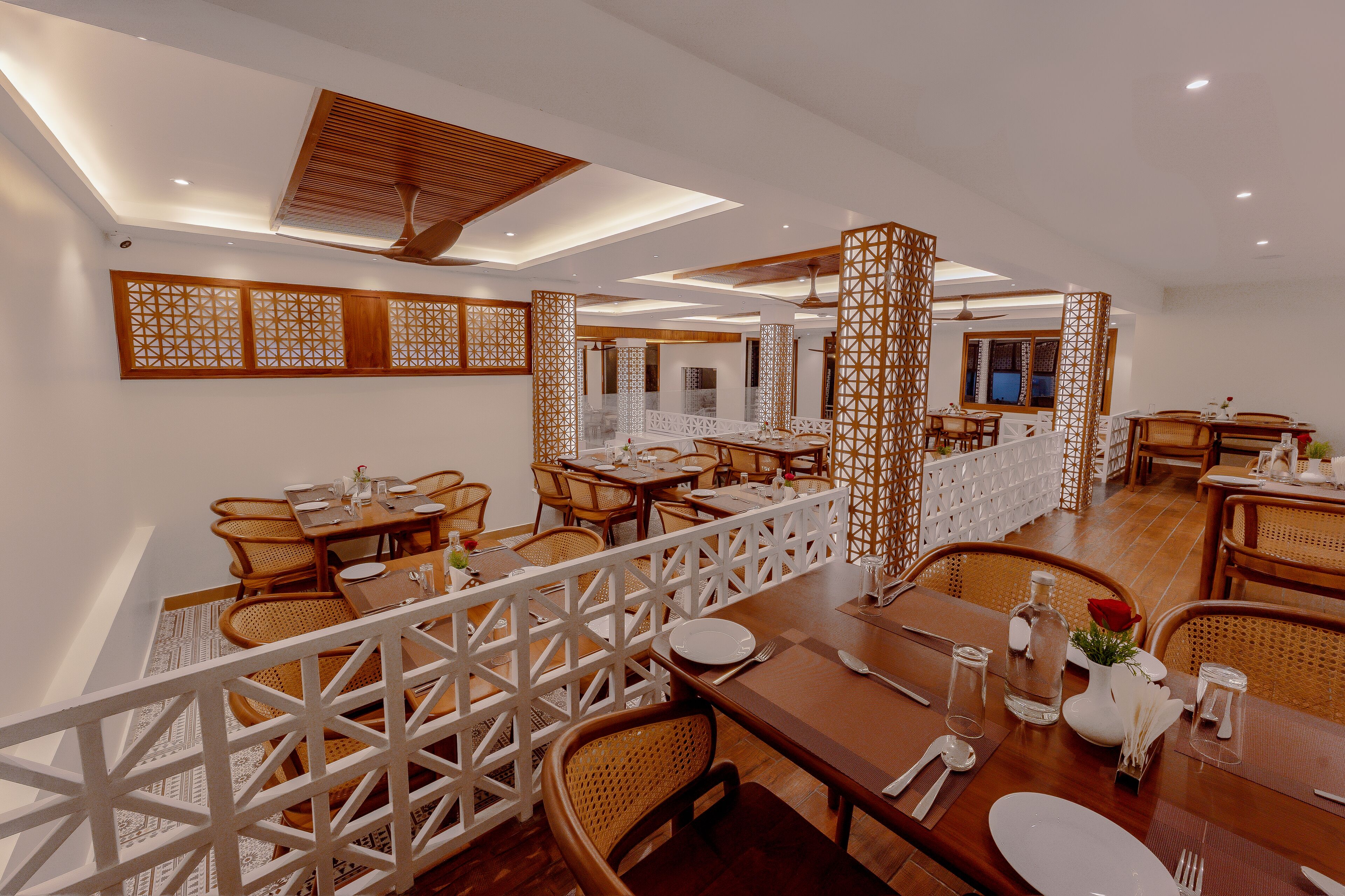 A restaurant with multiple dining tables, wicker chairs, and a white decorative partition.