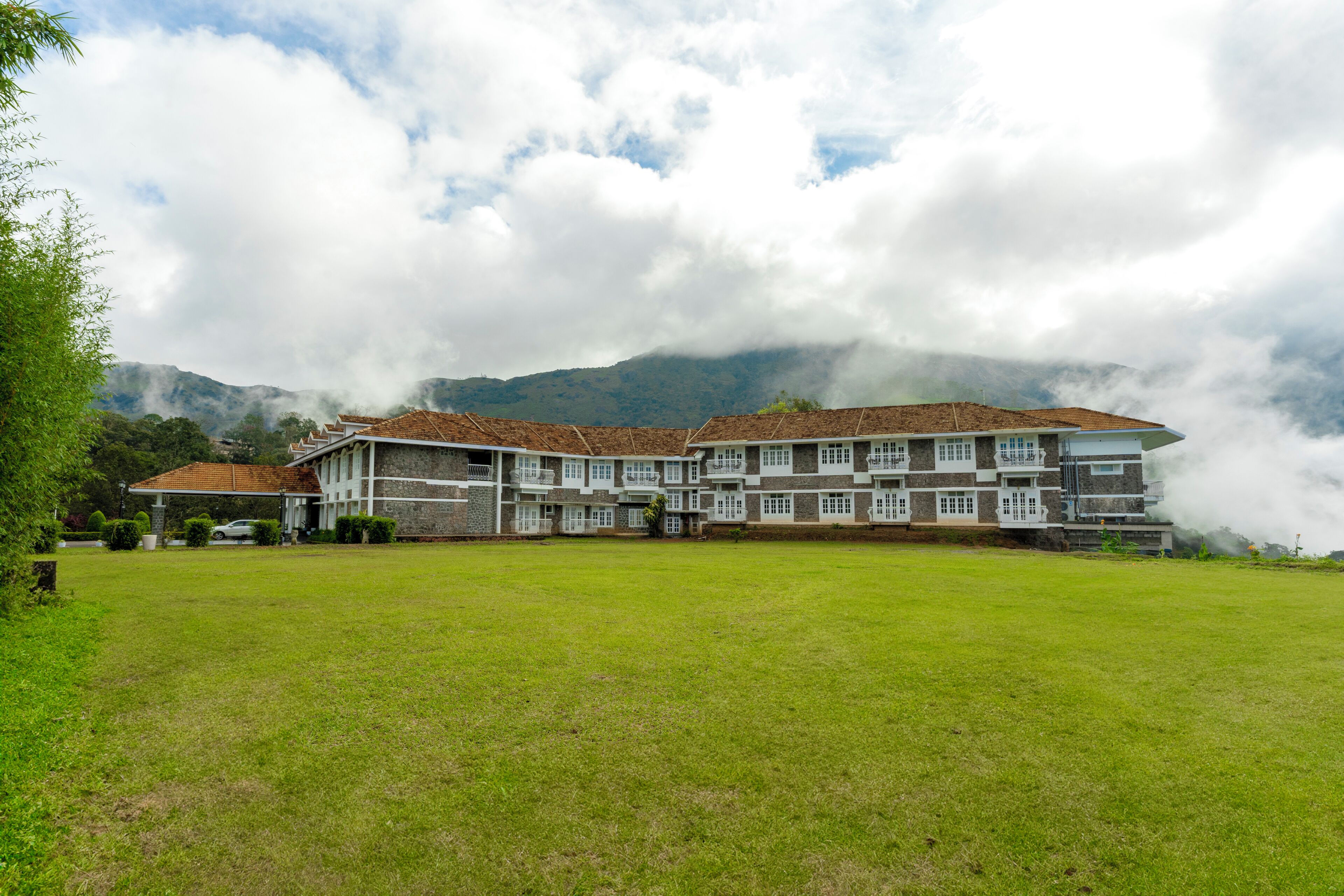 A large, multi-story building with a flat roof and multiple balconies, surrounded by a grassy area and mountains in the background.