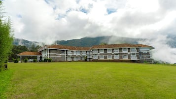 A large, multi-story building with a flat roof and multiple balconies, surrounded by a grassy area and mountains in the background.