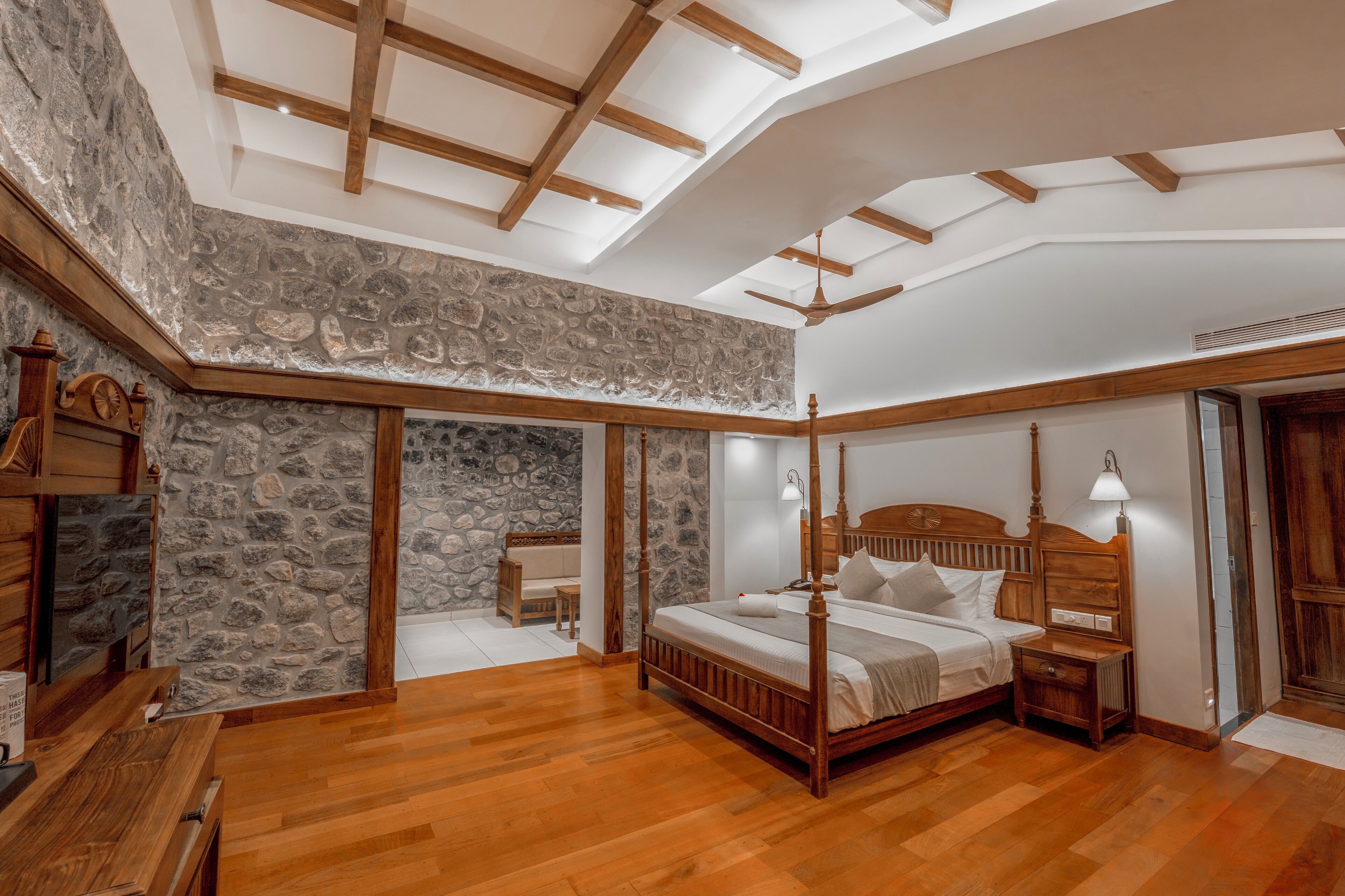 Un dormitorio con pared de piedra, vigas de madera en el techo y una cama con dosel, junto a una mesita de noche con lámpara.