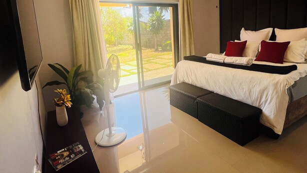 Ein modernes Schlafzimmer mit einem großen Bett, einem kleinen Tisch mit einer Vase und Blick auf einen Garten durch das Fenster.