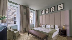Habitación de hotel con una cama grande, un banco, un escritorio y una silla. Hay cortinas, una ventana e imágenes enmarcadas en la pared.
