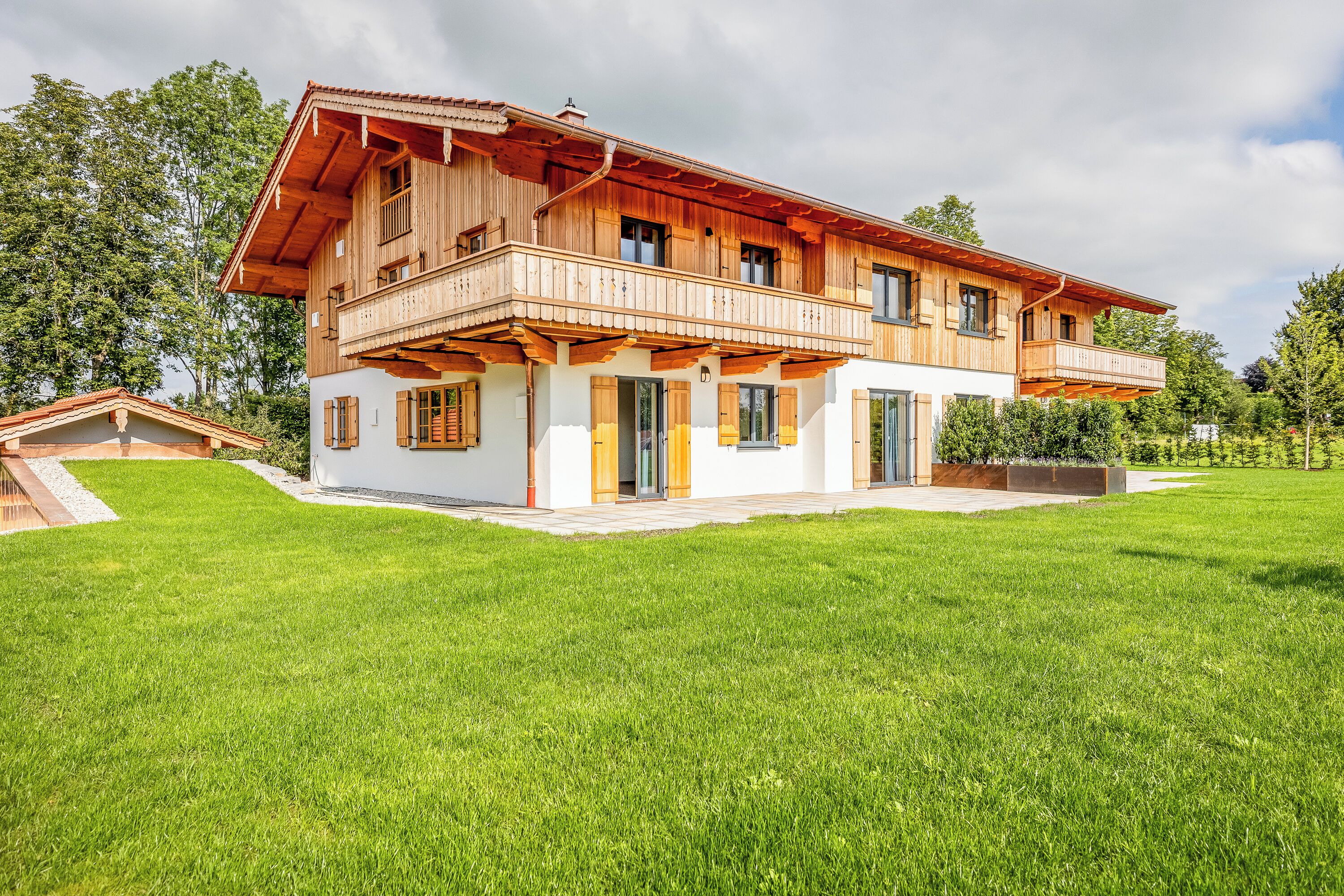 Una casa de madera de dos pisos con balcón, rodeada por un césped cuidado y árboles.