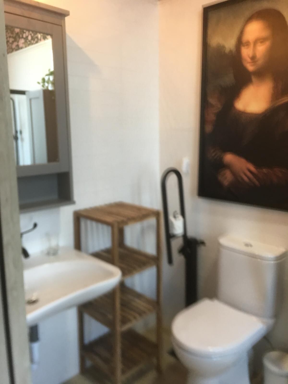 A bathroom with a white sink, wooden shelf, toilet, and a painting of the Mona Lisa on the wall.