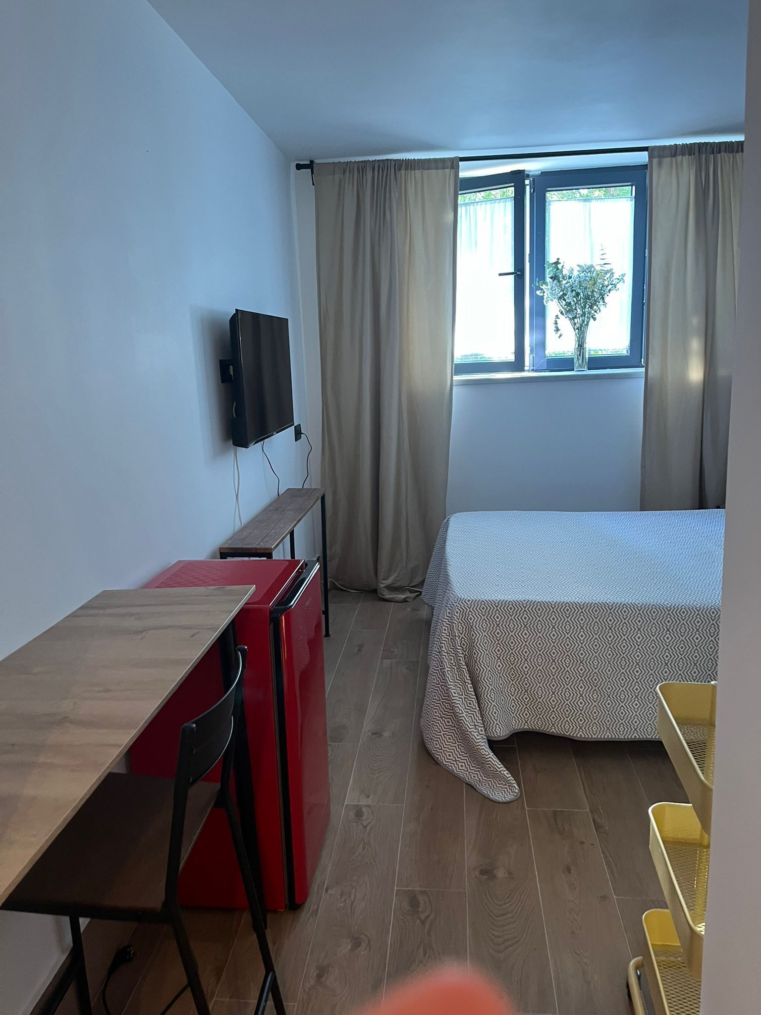 A compact room with a bed, a desk with a chair, a TV mounted on the wall, and a window with curtains.