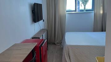 A compact room with a bed, a desk with a chair, a TV mounted on the wall, and a window with curtains.
