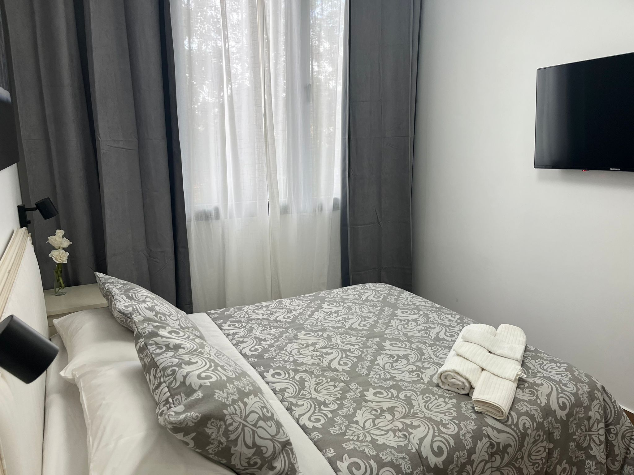 A bedroom with a bed, a TV on the wall, and a window with curtains.