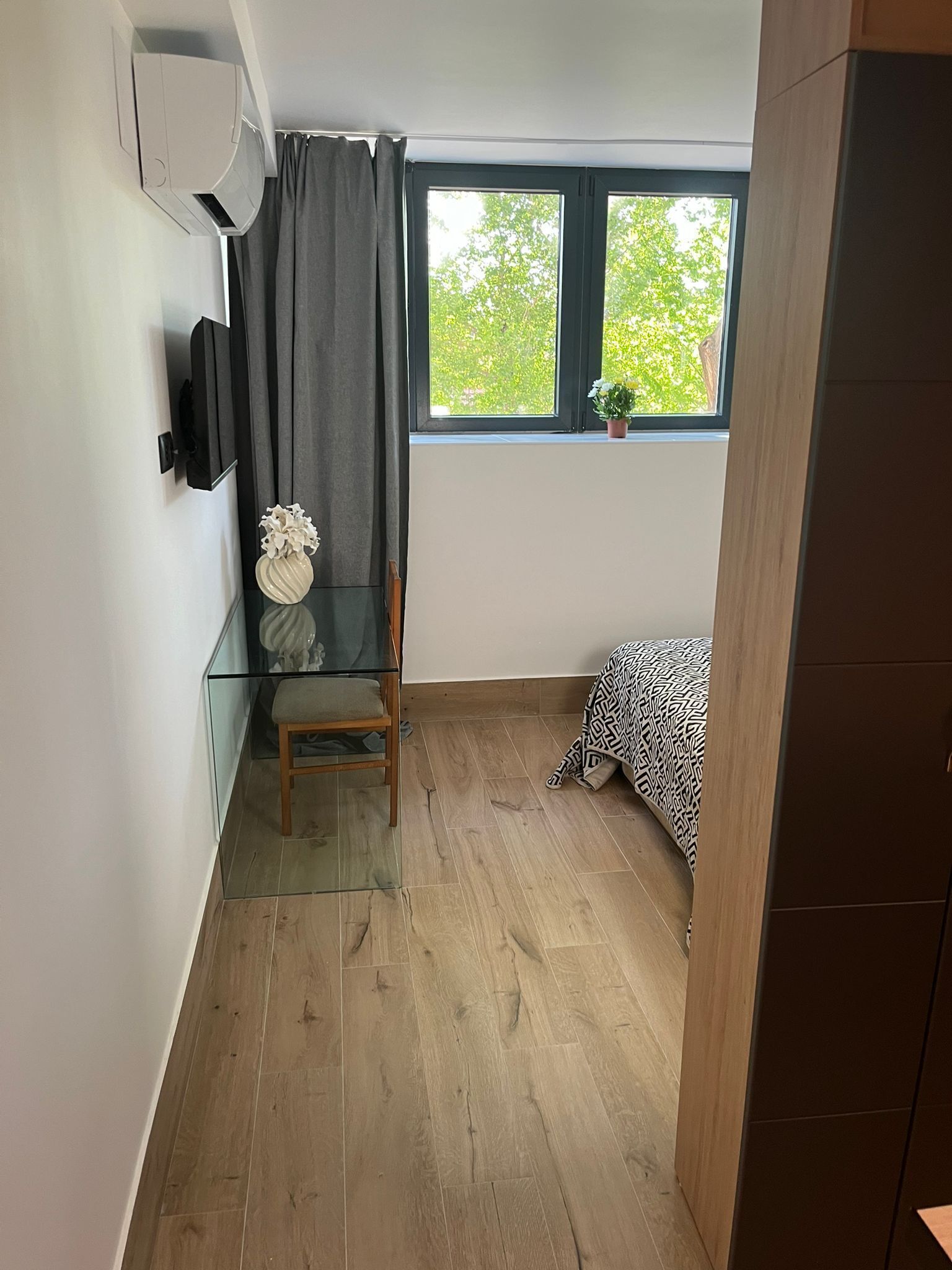 Una habitación pequeña con piso de madera, una cama con colcha estampada, una silla, una mesita con un florero, una ventana con vista a árboles y un equipo de aire acondicionado montado en la pared.
